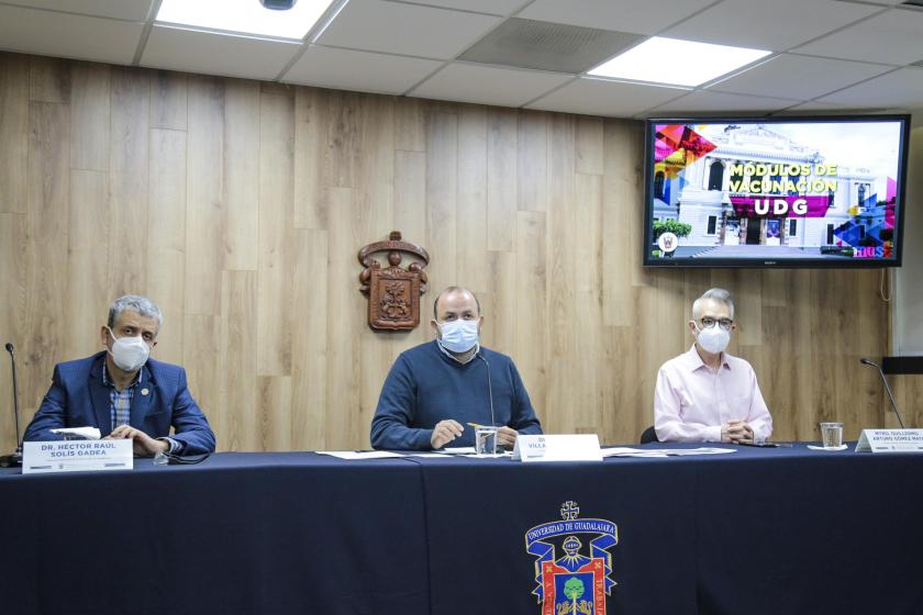 Tres personas sentadas. Frente a ellas un escritorio. En el fondo el emblema de la UdeG y un televisor en el que se lee "MÓDULOS DE VACUNACIÓN UDG".