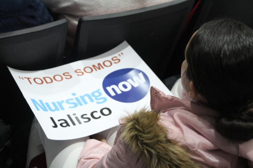 Una persona sentada sosteniendo un cartel sobre sus rodillas en las que se lee "TODOS SOMOS Nursing now Jalisco".