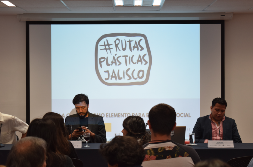 Dos personas sentadas. Detrás de ellas se proyecta una diapositiva en la que se lee "#RUTAS PLÁSICAS JALISCO".