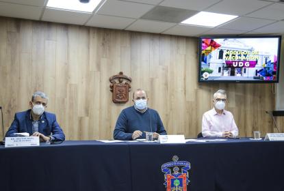 Tres personas sentadas. Frente a ellas un escritorio. En el fondo el emblema de la UdeG y un televisor en el que se lee "MÓDULOS DE VACUNACIÓN UDG".