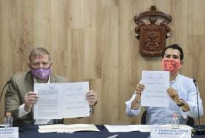 Dos autoridades mostrando papeles a la cámara. En el fondo el escudo de la UdeG.