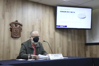 Una persona sentada, frente a él un micrófono. En el fondo la insignia de la UdeG y un televisor.